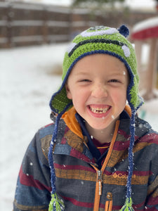 Smiling young boy wearing a crochet monster beanie. In the background it's snowing.